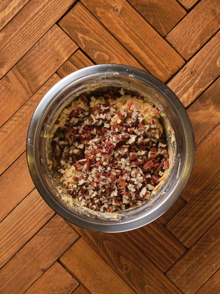 A metal bowl with cookie dough mixed with chopped pecans on a wooden herringbone floor.