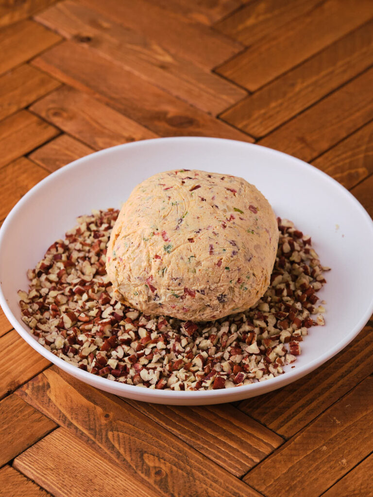 A ball of cheese covered with herbs and dried fruit is placed in a white bowl filled with chopped nuts.