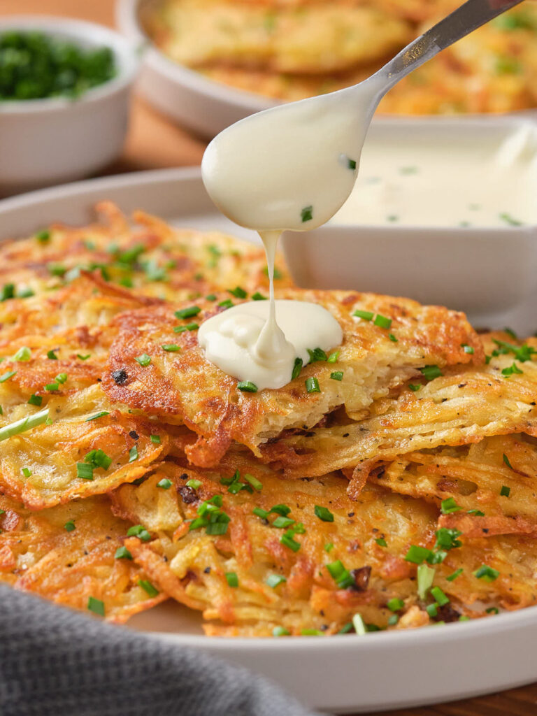 A pile of golden crispy potato fritters with chopped chives being drizzled with sour cream.