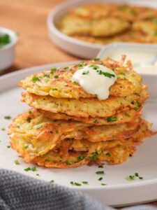 A stack of potato fritters topped with sour cream and garnished with chopped chives on a white plate.