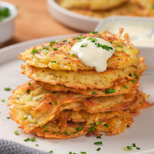 A stack of potato fritters topped with sour cream and garnished with chopped chives on a white plate.