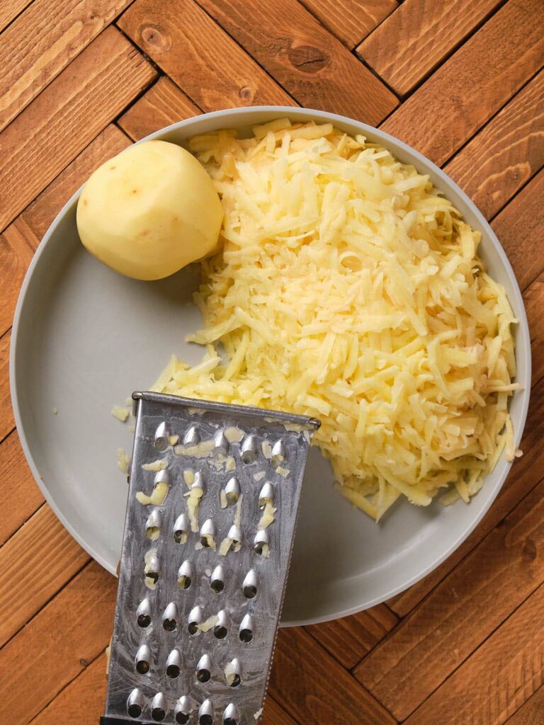 A peeled potato and a pile of grated potato sit on a gray plate with a metal grater.