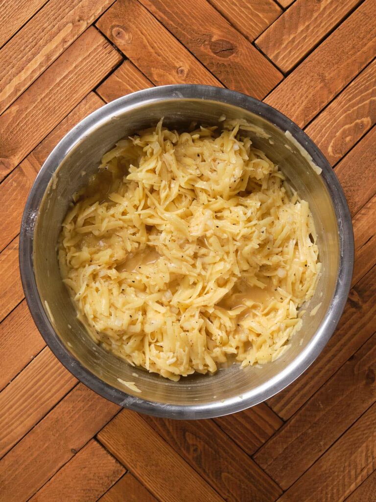 A metal bowl containing grated potatoes mixed with herbs, placed on a wooden surface.