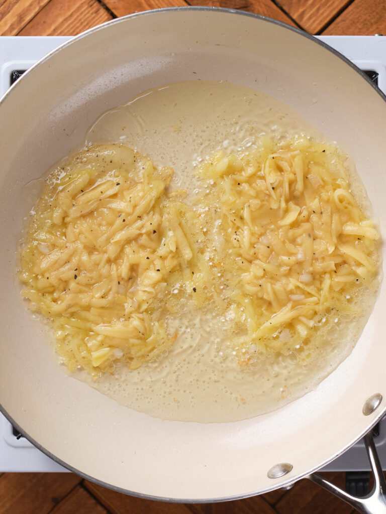 Grated potatoes frying in a pan with oil on a stove.