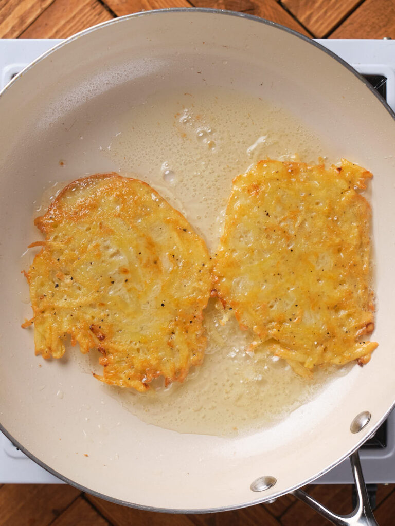 Two potato fritters frying in a non-stick pan with a light coating of oil.