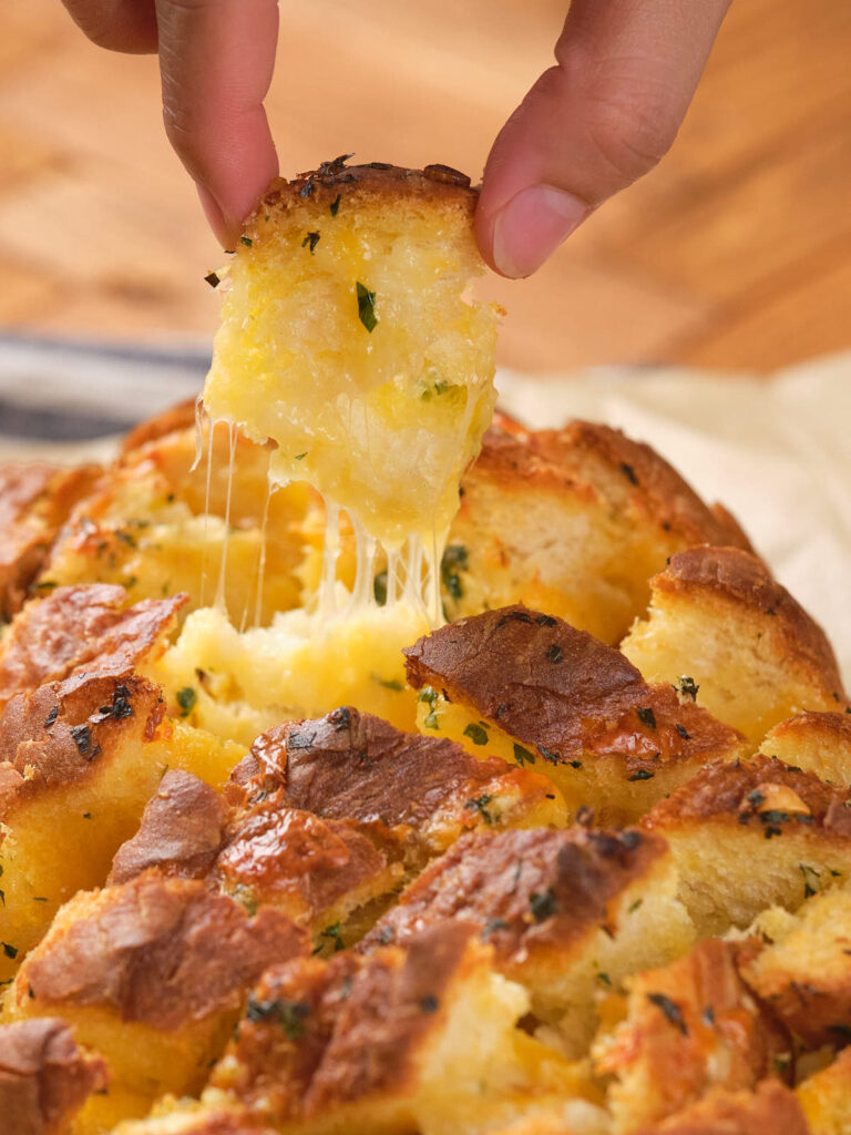 Hand pulling a piece of garlic cheese bread from a loaf, with melted cheese stretching.