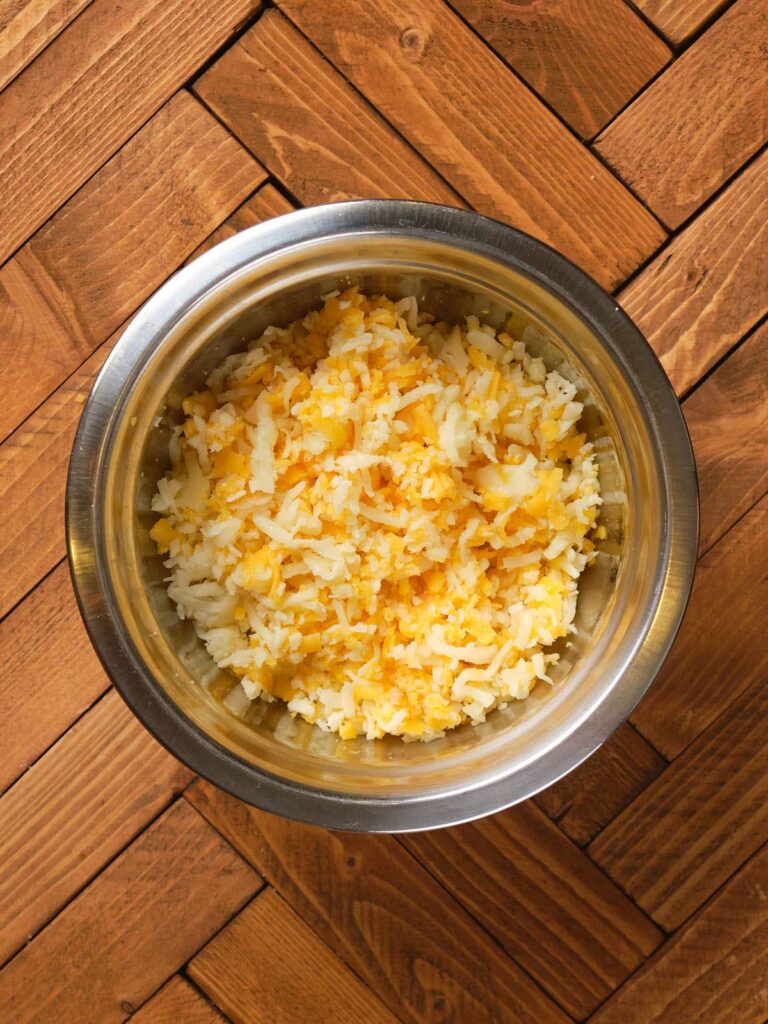 Bowl of shredded yellow and white cheese on a wooden herringbone-patterned surface.