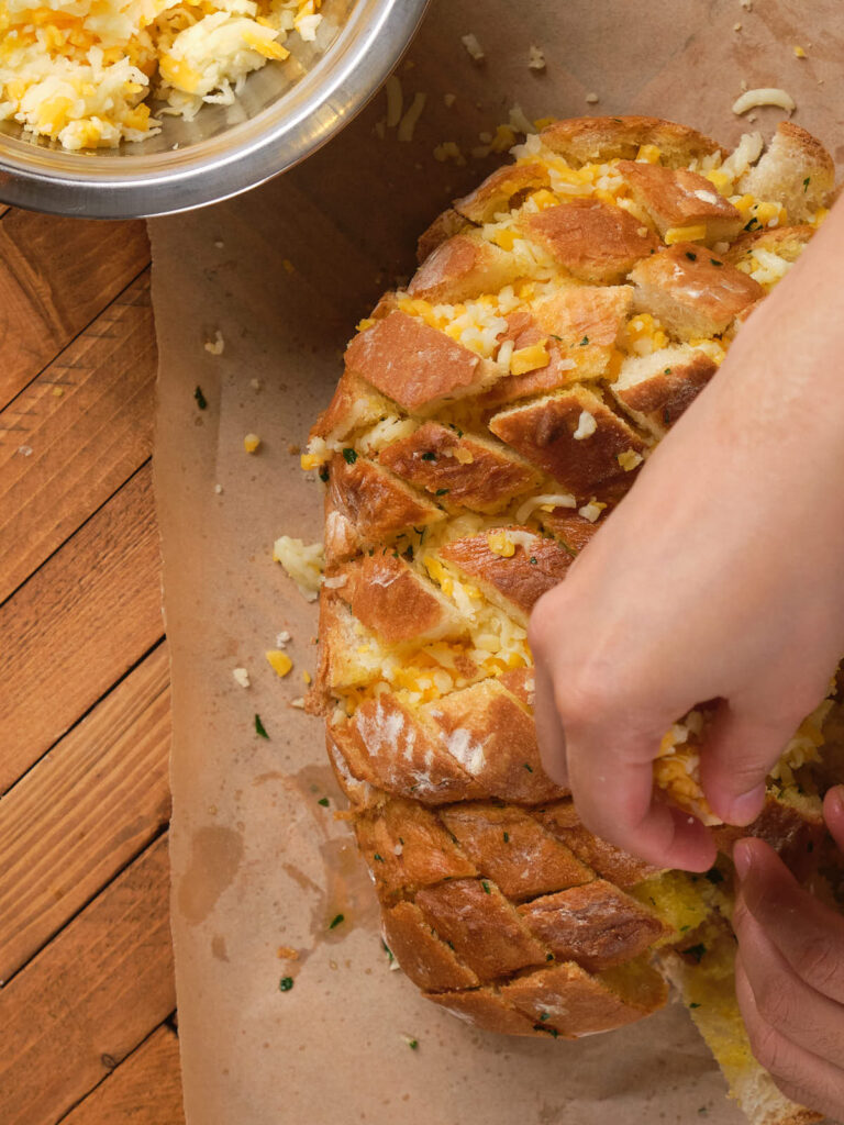 Hands preparing garlic cheese bread on a wooden surface with a bowl of shredded cheese nearby.