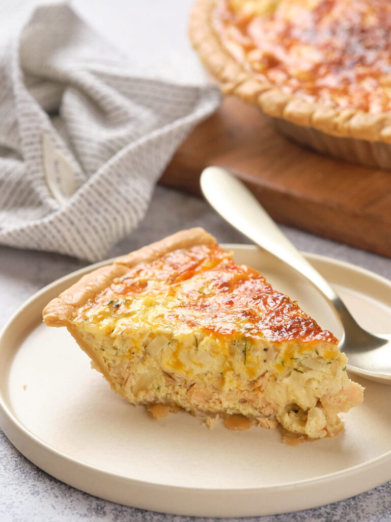 A slice of salmon quiche sits on a beige plate with a spoon beside it.