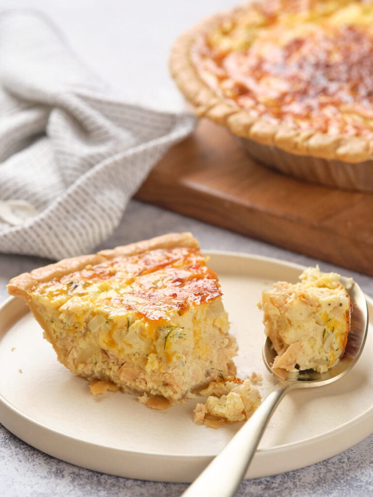 A slice of salmon quiche on a plate with a spoonful beside it.