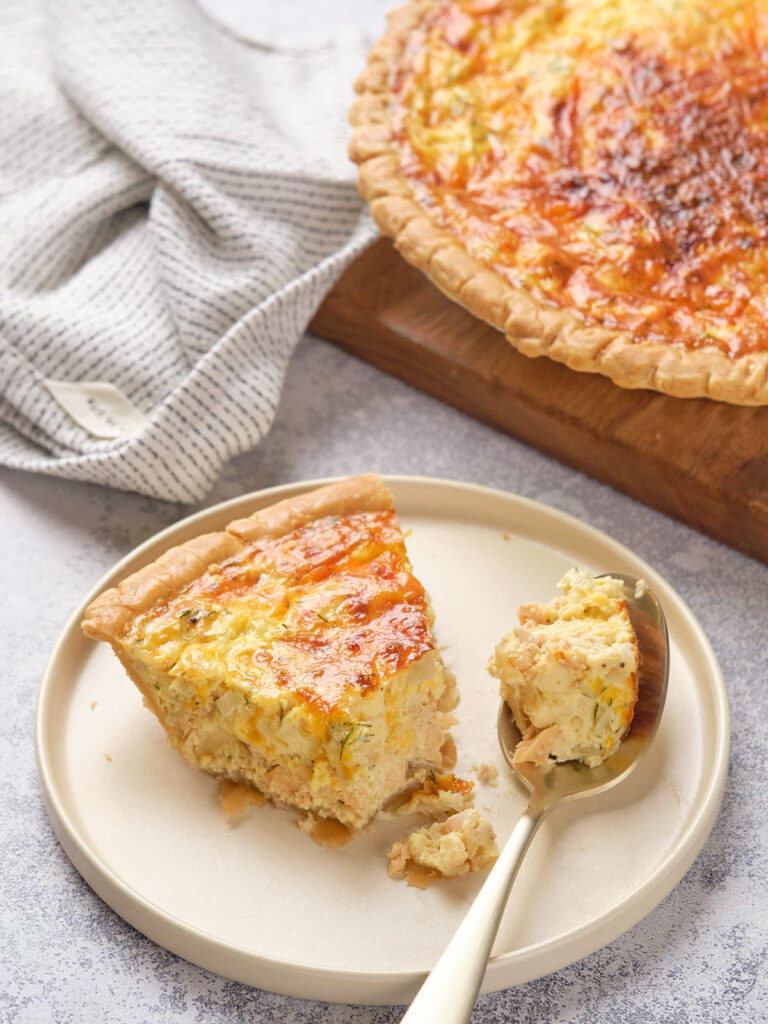 Slice of salmon quiche on a plate with a fork, next to a whole quiche on a wooden board.