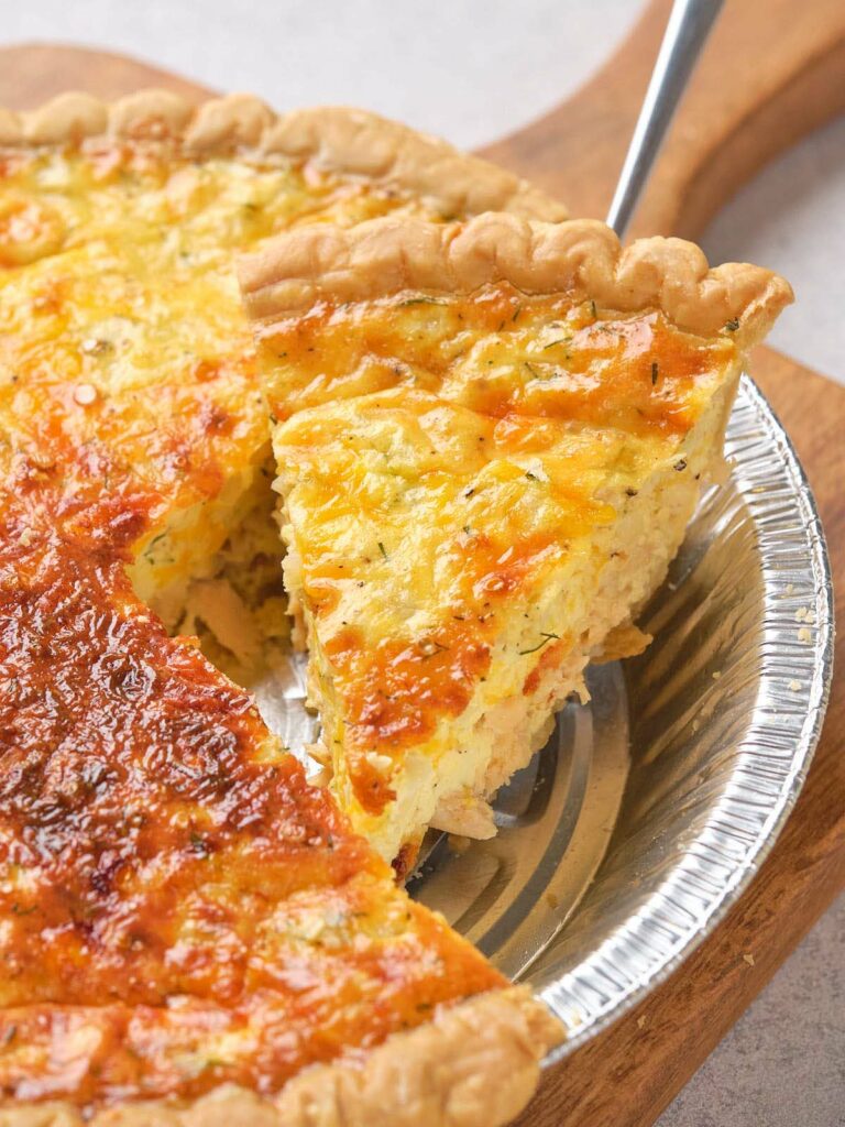 A slice of salmon quiche in a metal pie tin on a wooden board.