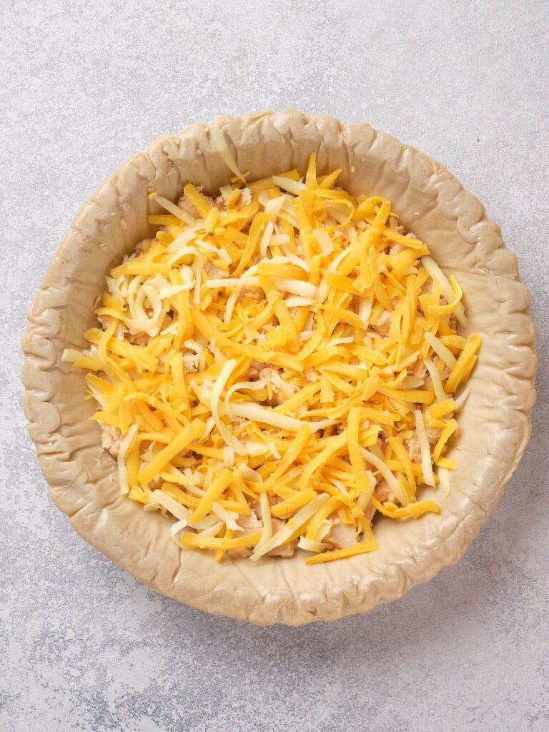 An unbaked pie crust filled with shredded yellow and white cheese on a light gray surface.