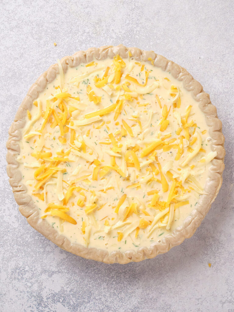 Unbaked quiche with shredded cheese and herbs in a pie crust on a light gray surface.