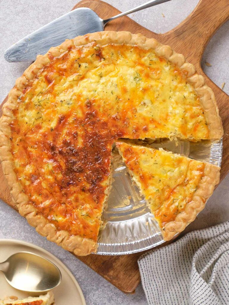 A baked quiche with a golden crust, one slice removed, on a wooden board.