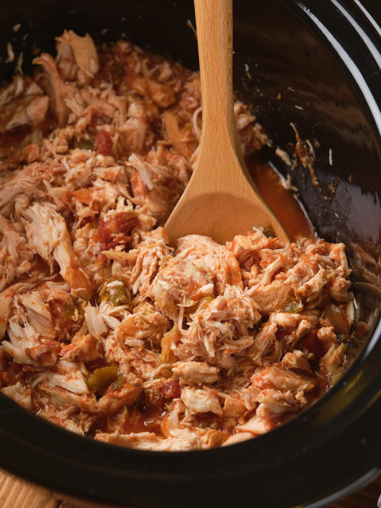 Shredded chicken with tomatoes and peppers in a slow cooker, stirred with a wooden spoon.