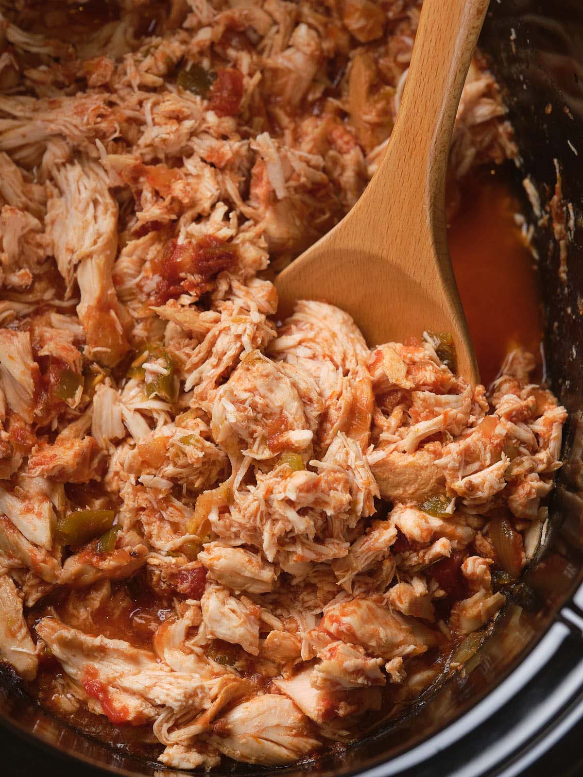 Shredded chicken with tomatoes and green peppers being stirred with a wooden spoon.