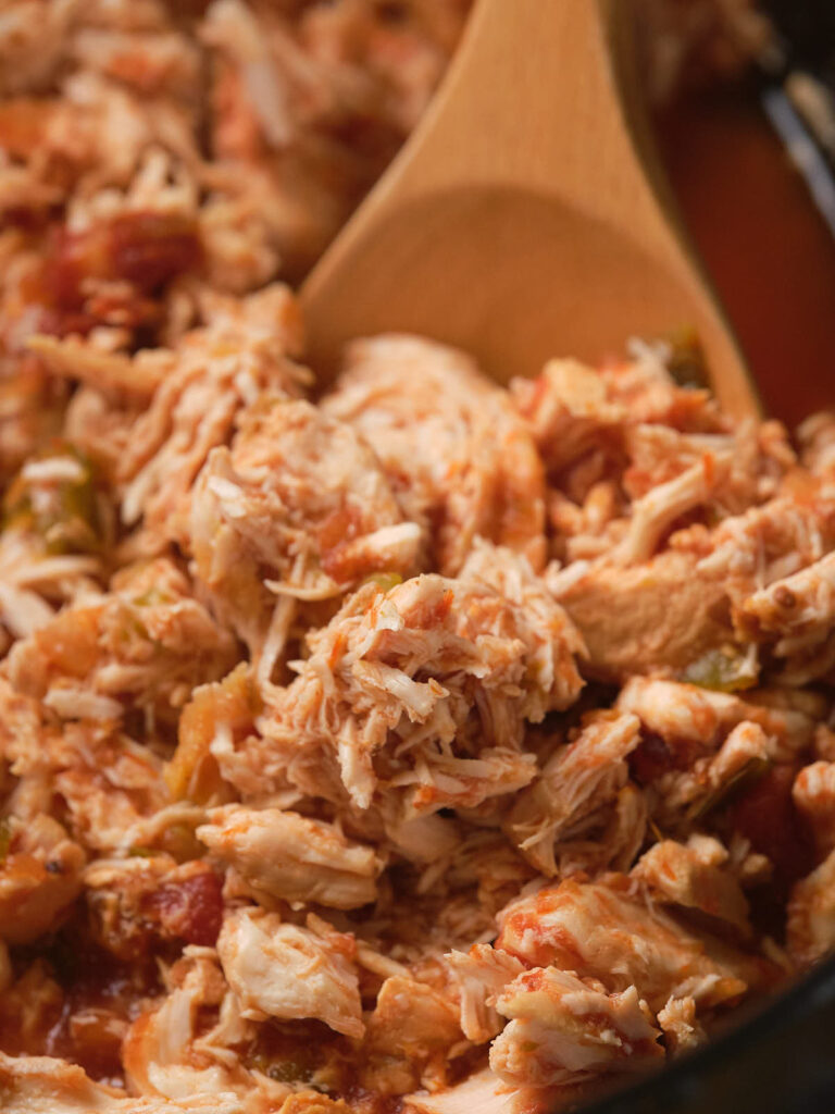 Close-up of shredded chicken mixed with diced tomatoes and green peppers in a wooden spoon.