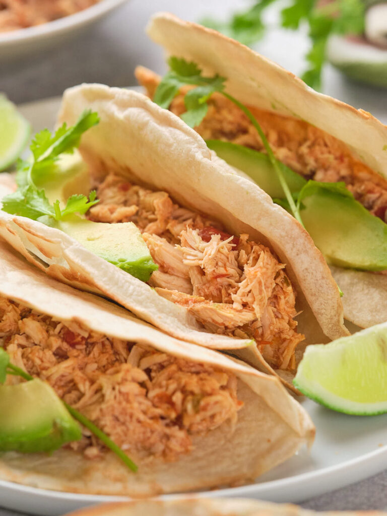 Three tacos filled with shredded chicken and avocado slices, garnished.