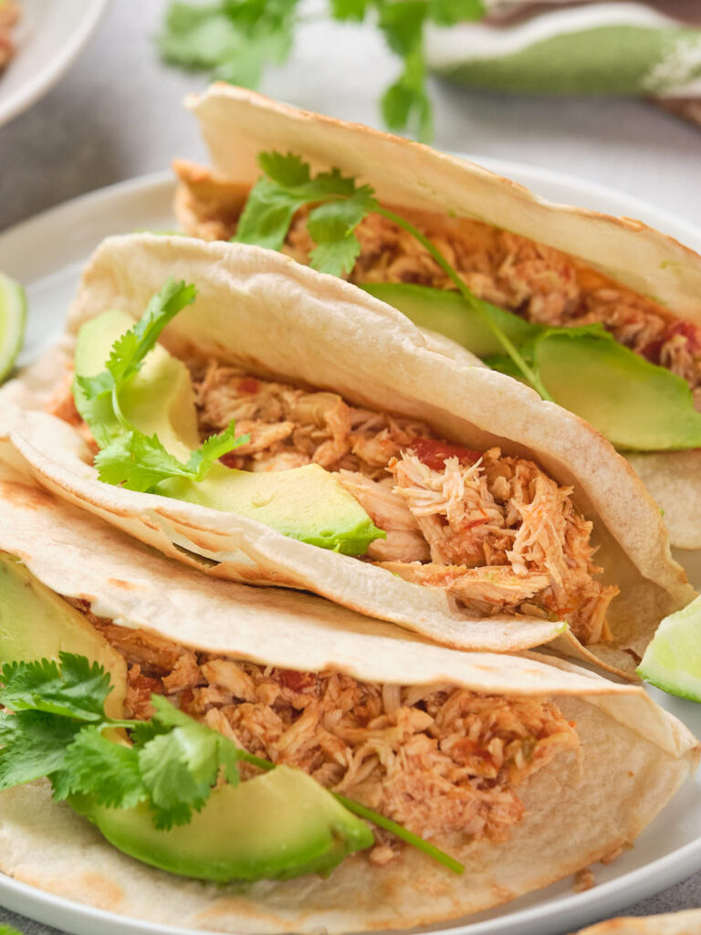 Three tacos filled with shredded chicken, avocado slices, and cilantro on a plate.