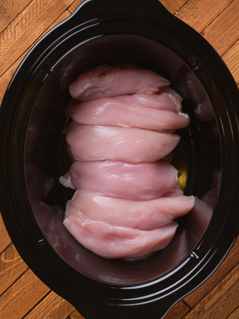 Raw chicken breasts placed in a black crockpot on a wooden surface.