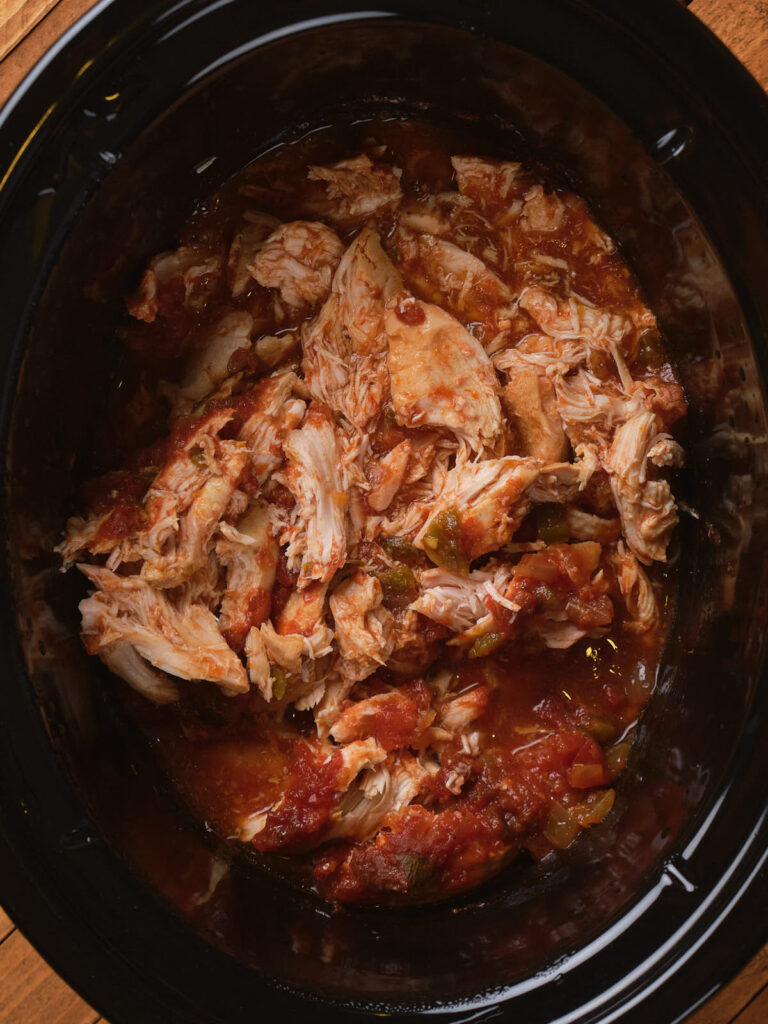 Shredded chicken stew in a black slow cooker, mixed with diced tomatoes and peppers.