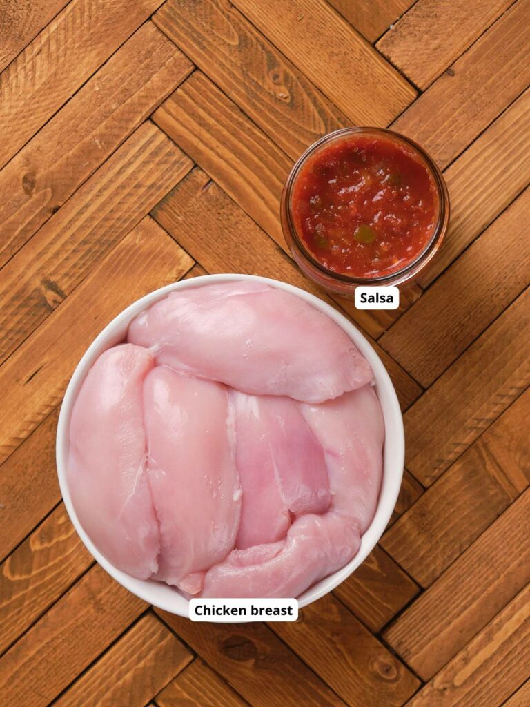 A bowl of raw chicken breast on a wooden surface next to a small jar of salsa.