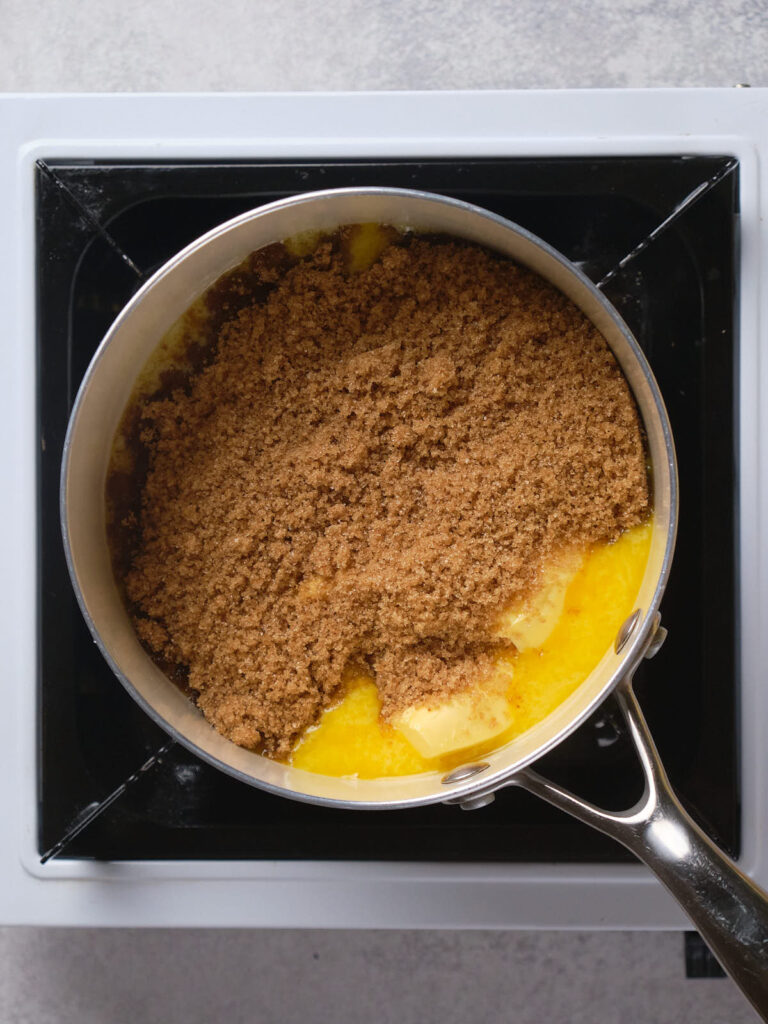 A saucepan on a stove contains partially melted butter and brown sugar.