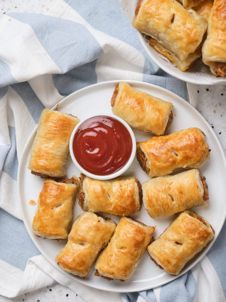 A plate of golden-brown sausage rolls with a small bowl of ketchup at the center.