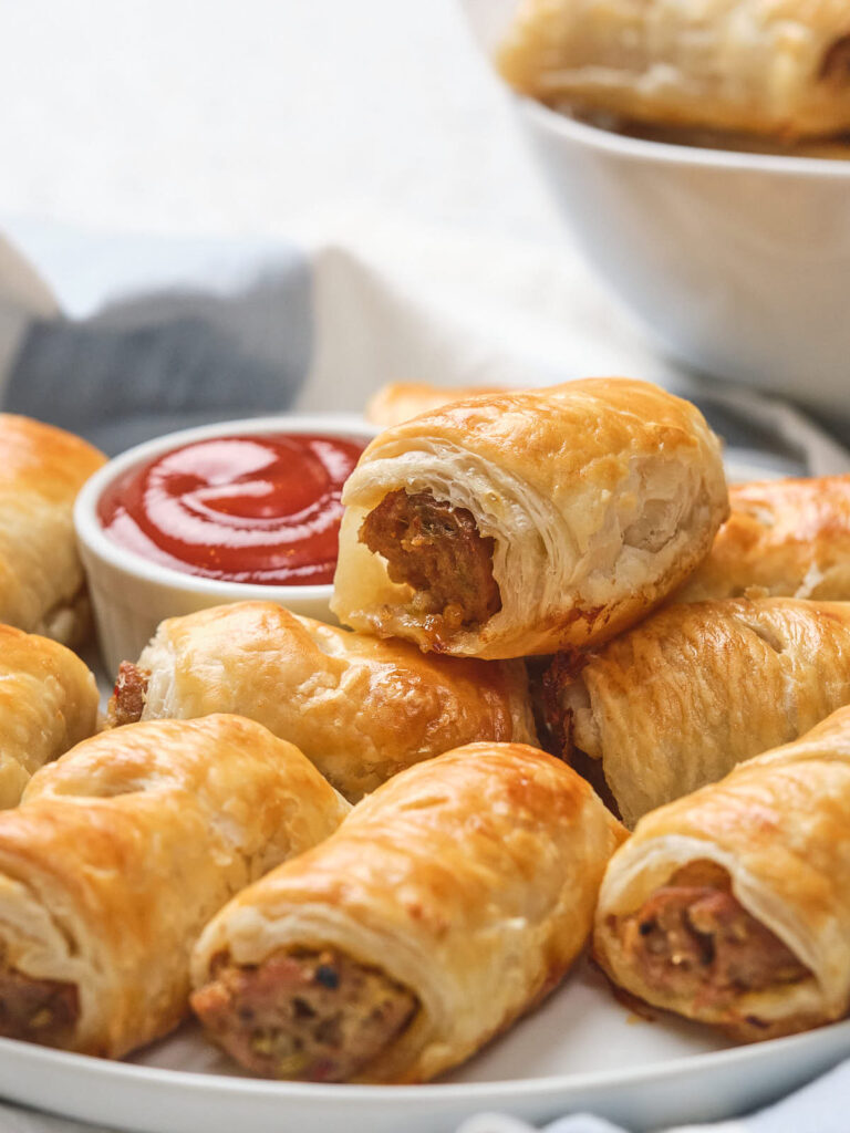 A plate of golden-brown sausage rolls with one partially eaten, served with a small bowl of ketchup.