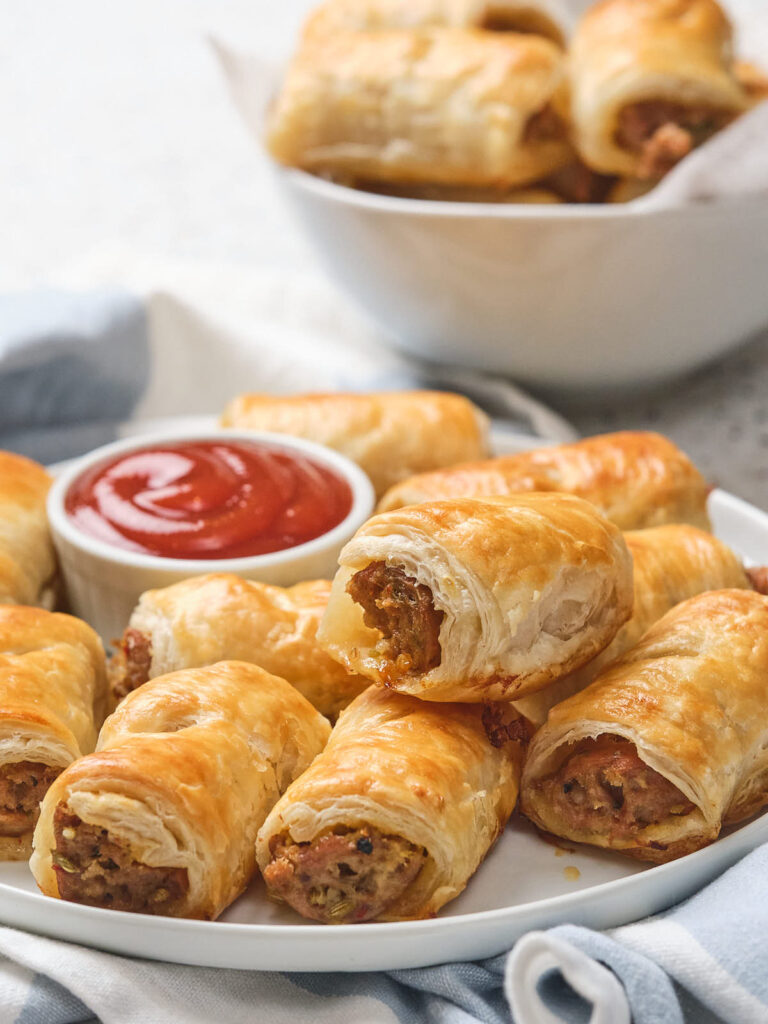 A plate of golden-brown sausage rolls with a small bowl of tomato sauce.