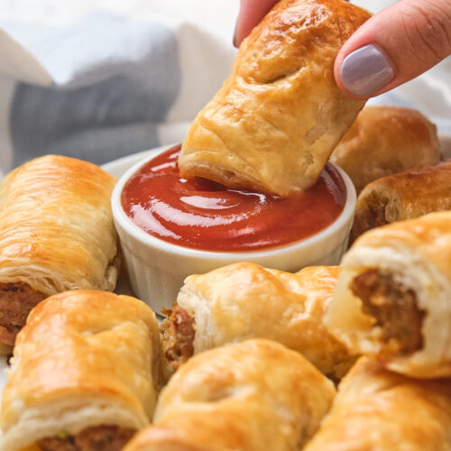 A hand dips a sausage roll into a small bowl of ketchup.