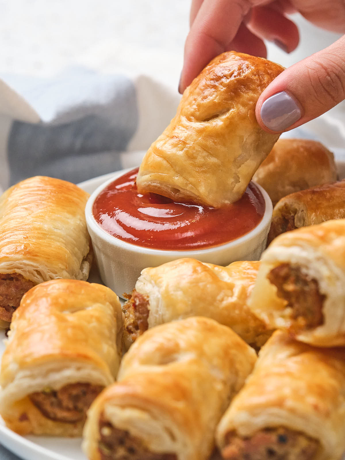 A hand dips a sausage roll into a small bowl of ketchup.