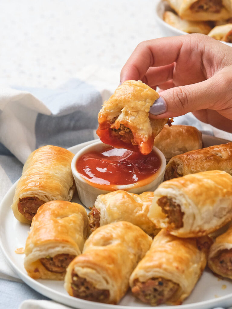 A hand dipping a sausage roll into red sauce on a plate filled with more sausage rolls.