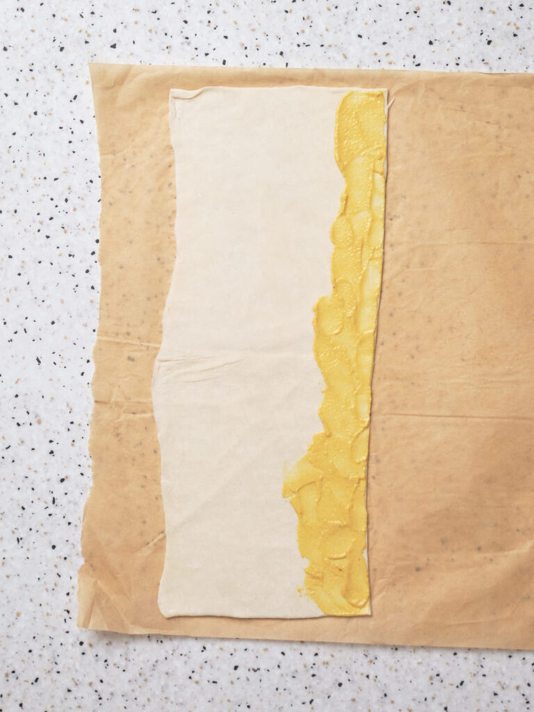 A sheet of pastry partly covered with a layer of mustard, placed on parchment paper.