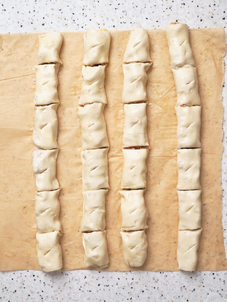 Four rows of uncooked pastry rolls, each cut into evenly-sized pieces, placed on brown parchment paper on a speckled countertop.