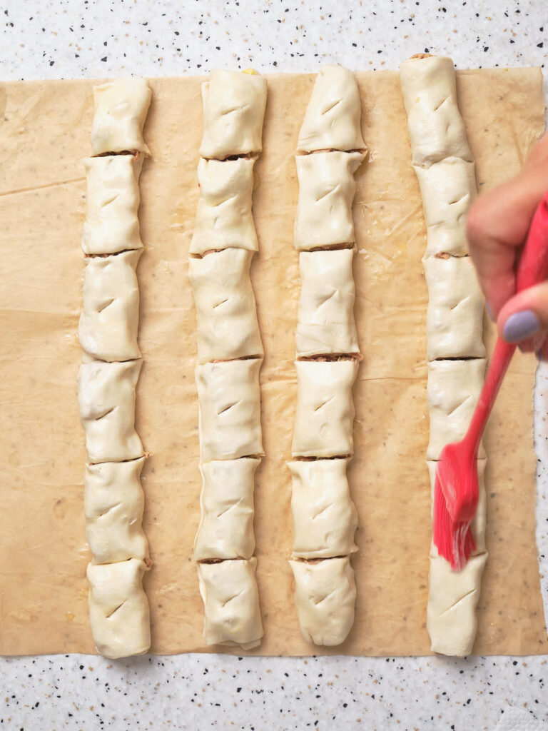 Four rows of unbaked pastry pieces on parchment paper, with a hand brushing one row using a red brush.