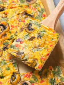 A slice of vegetable frittata with mushrooms, bell peppers, and spinach on a wooden spatula.