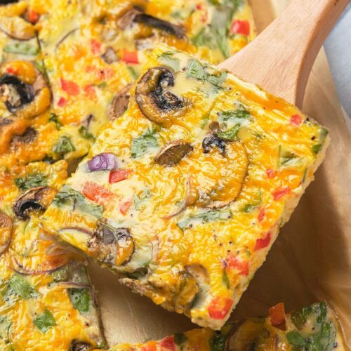 A slice of vegetable frittata with mushrooms, bell peppers, and spinach on a wooden spatula.