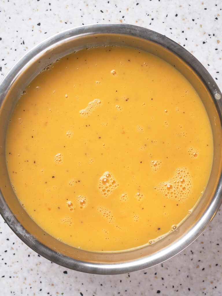 A stainless steel bowl filled with whisked eggs, placed on a speckled white countertop.