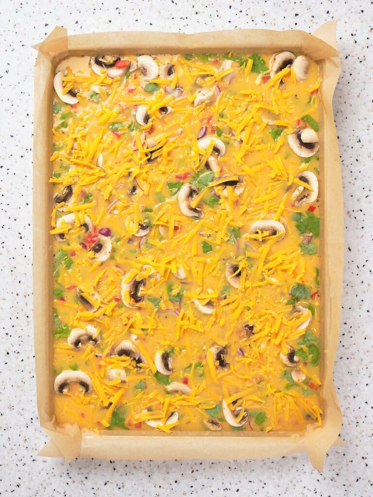 A baking sheet with a raw omelette mixture topped with mushrooms, vegetables, and shredded cheddar cheese.