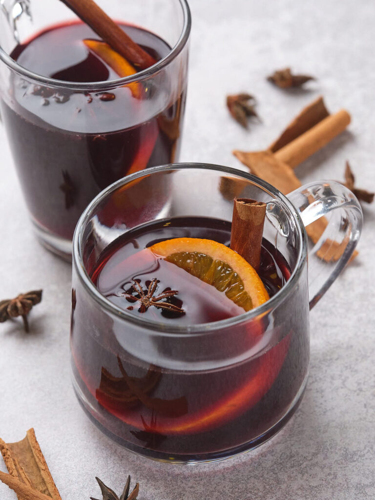 Two glass mugs of mulled wine with orange slices, cinnamon sticks, and star anise.