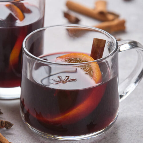 A clear mug filled with dark red mulled wine, garnished with an orange slice, a cinnamon stick, and star anise.