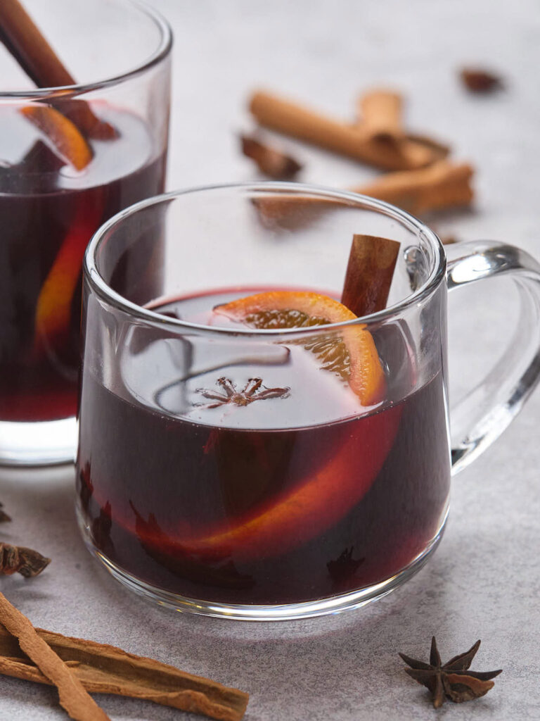 A clear mug filled with dark red mulled wine, garnished with an orange slice, a cinnamon stick, and star anise.