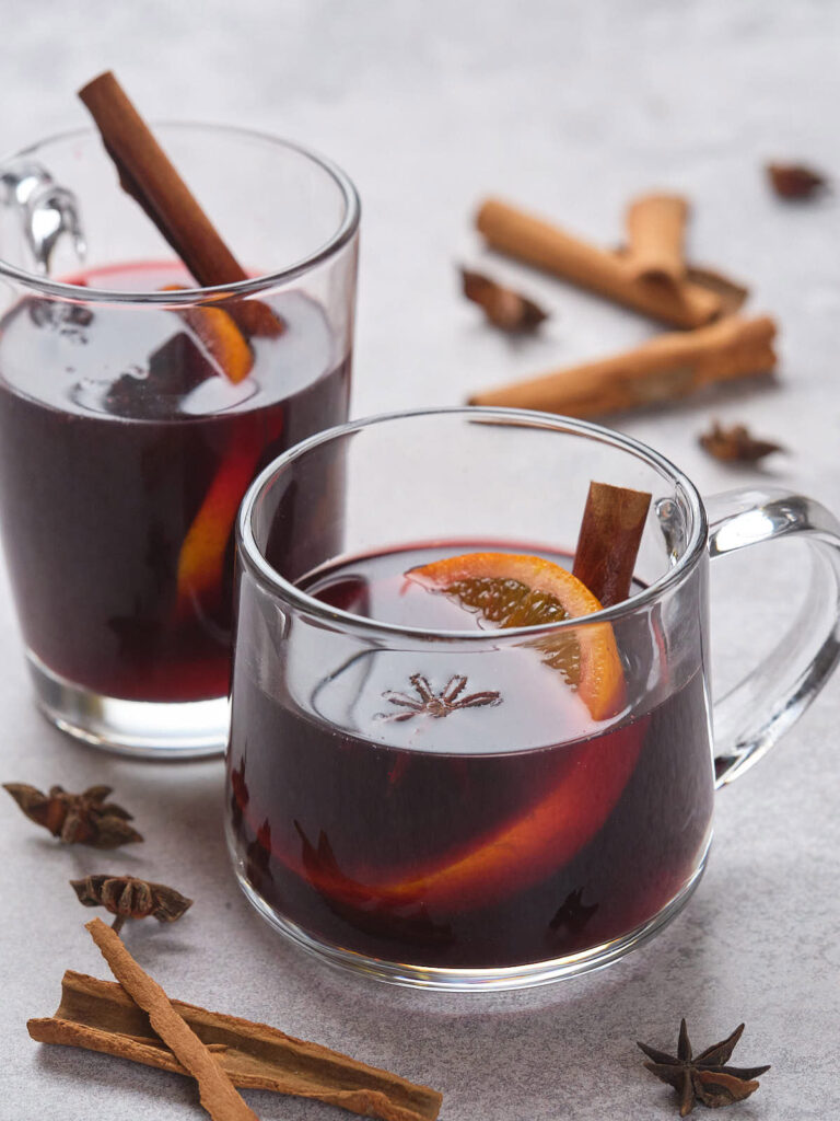 Two clear mugs of mulled wine with orange slices and cinnamon sticks on a gray surface.