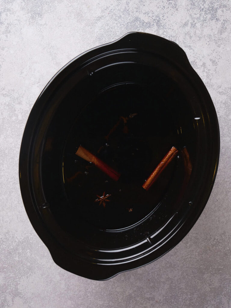 A black slow cooker with dark liquid, cinnamon sticks, and a star anise inside.