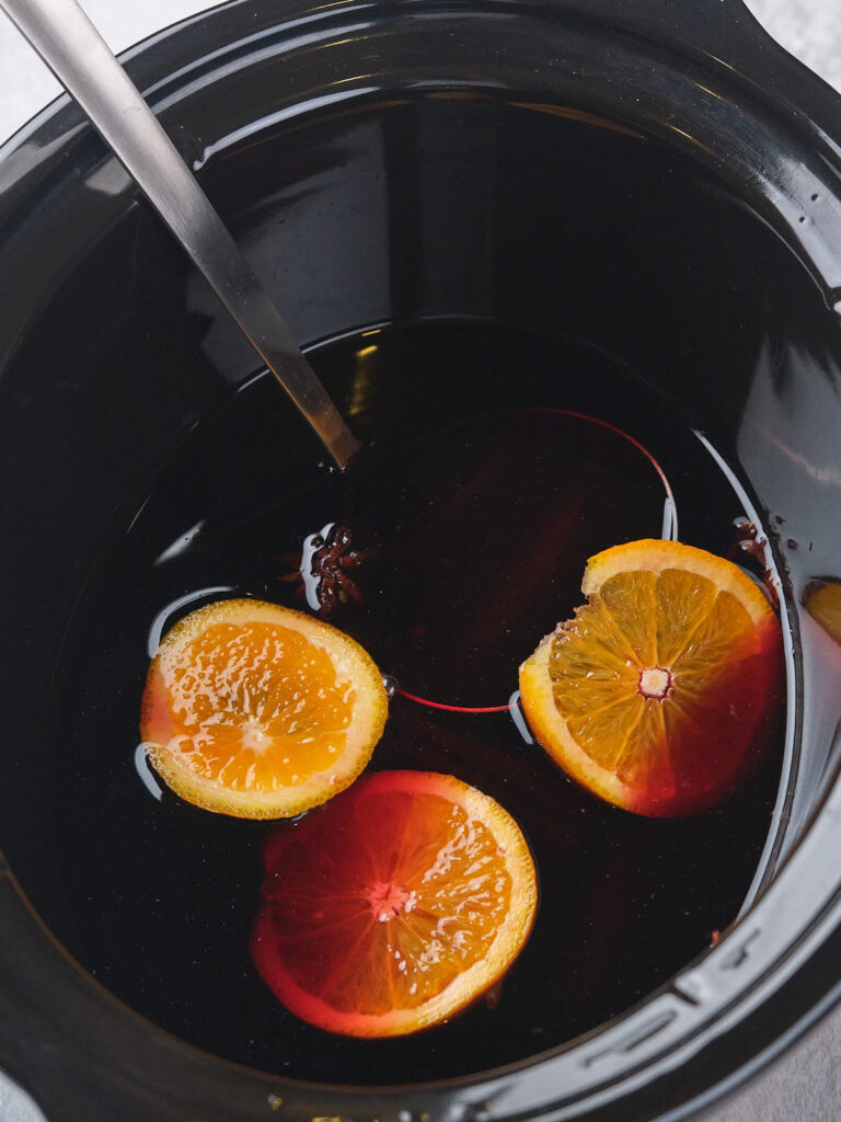 A black slow cooker containing a dark liquid with three slices of orange floating on top.