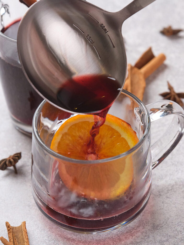 A ladle pours mulled wine into a glass mug containing an orange slice.