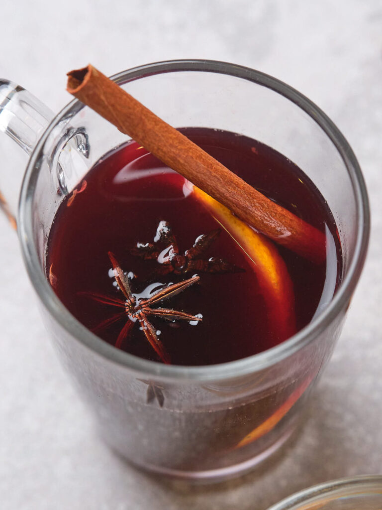 A glass mug of mulled wine garnished with a cinnamon stick, a slice of orange, and star anise.