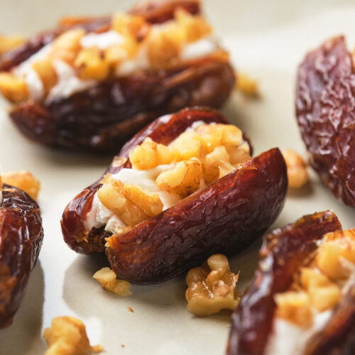 Close-up of dates stuffed with a creamy filling and topped with chopped nuts on a light-colored surface.